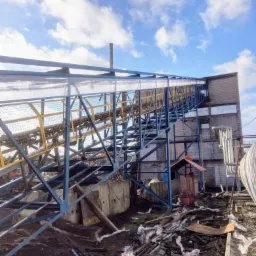 Konstrukcja stalowa z żółtymi elementami prowadząca do budynku z cegły, widok z dołu na tle błękitnego nieba z chmurami.