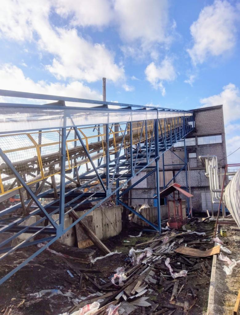 Konstrukcja stalowa z żółtymi elementami prowadząca do budynku z cegły, widok z dołu na tle błękitnego nieba z chmurami.