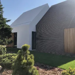 Nowoczesny dom z białymi i ciemnymi elewacjami, otoczony zielenią i niskimi krzewami iglastymi, oświetlony porannym słońcem.