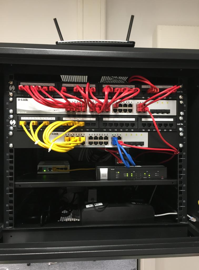 Widok z przodu szafy rack z zamontowanymi urządzeniami sieciowymi: switche D-Link i Cisco, routery oraz okablowanie w kolorach czerwonym, żółtym i niebieskim.