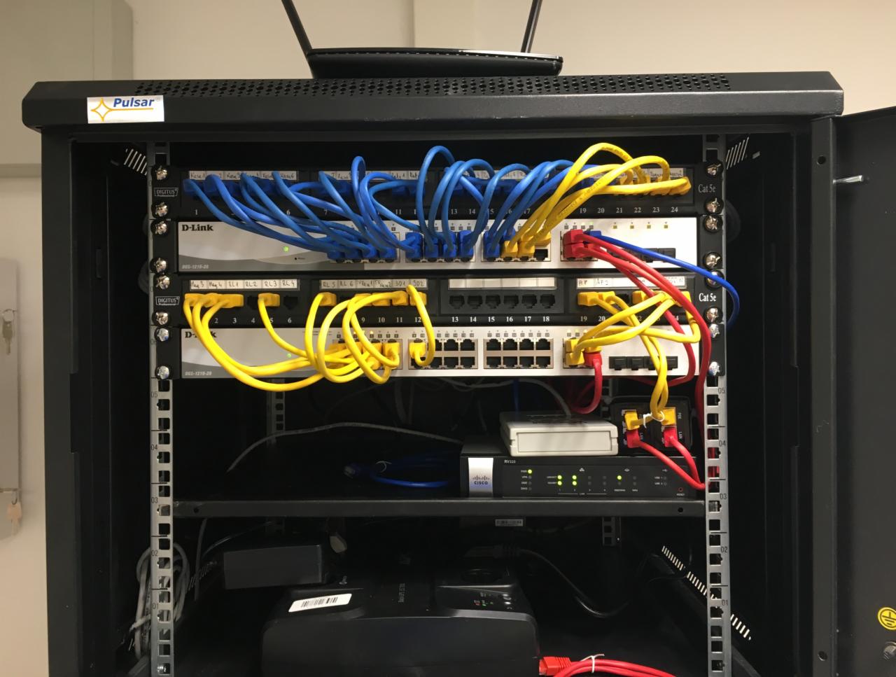 Szafa rack z zamontowanymi urządzeniami sieciowymi D-Link i Cisco, okablowanie Cat5e w kolorach niebieskim, żółtym i czerwonym, zasilacz UPS na dole szafy.