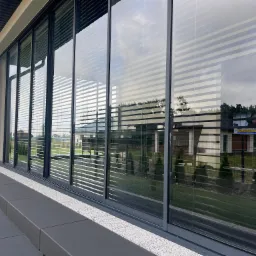 Przesuwne szklane drzwi balkonowe z szarymi ramami i poziomymi żaluzjami wewnętrznymi, widok z zewnątrz na taras i ogród.