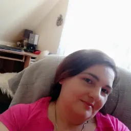 Uśmiechnięta kobieta w różowej koszulce siedzi na fotelu w jasnym pokoju na poddaszu. W tle widoczne biurko i okno z firanką. Dekoracja na ścianie.