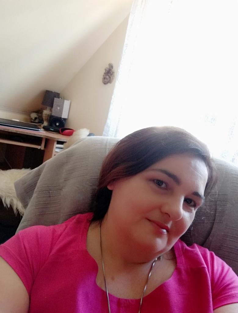 Uśmiechnięta kobieta w różowej koszulce siedzi na fotelu w jasnym pokoju na poddaszu. W tle widoczne biurko i okno z firanką. Dekoracja na ścianie.