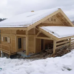 Surowy, drewniany dom w górach, częściowo pokryty śniegiem, z widocznymi elementami konstrukcyjnymi i stertą drewna opałowego.