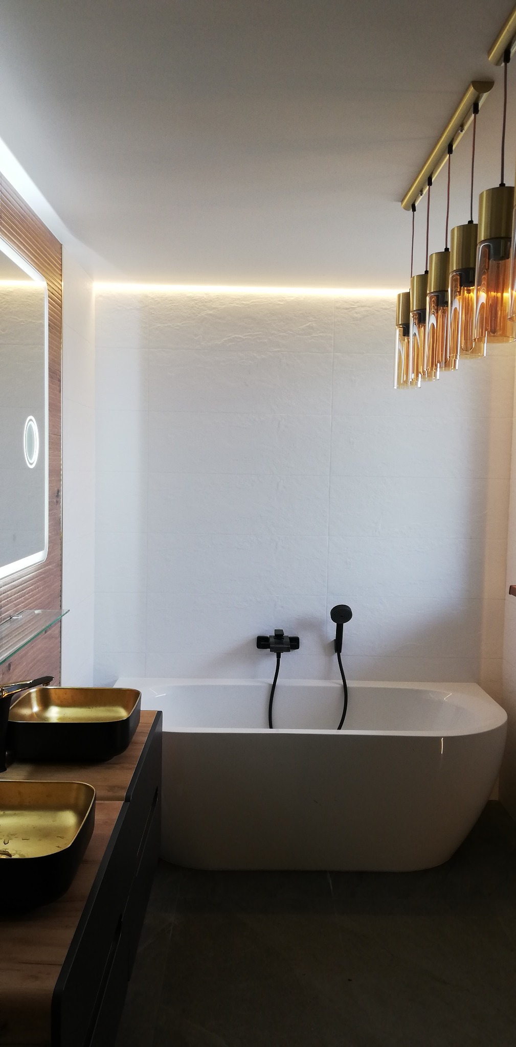 Nowoczesna łazienka z białą wanną, czarną armaturą, złotymi umywalkami nablatowymi i oświetleniem LED podsufitowym oraz wiszącymi lampami z elementami szkła i złota.