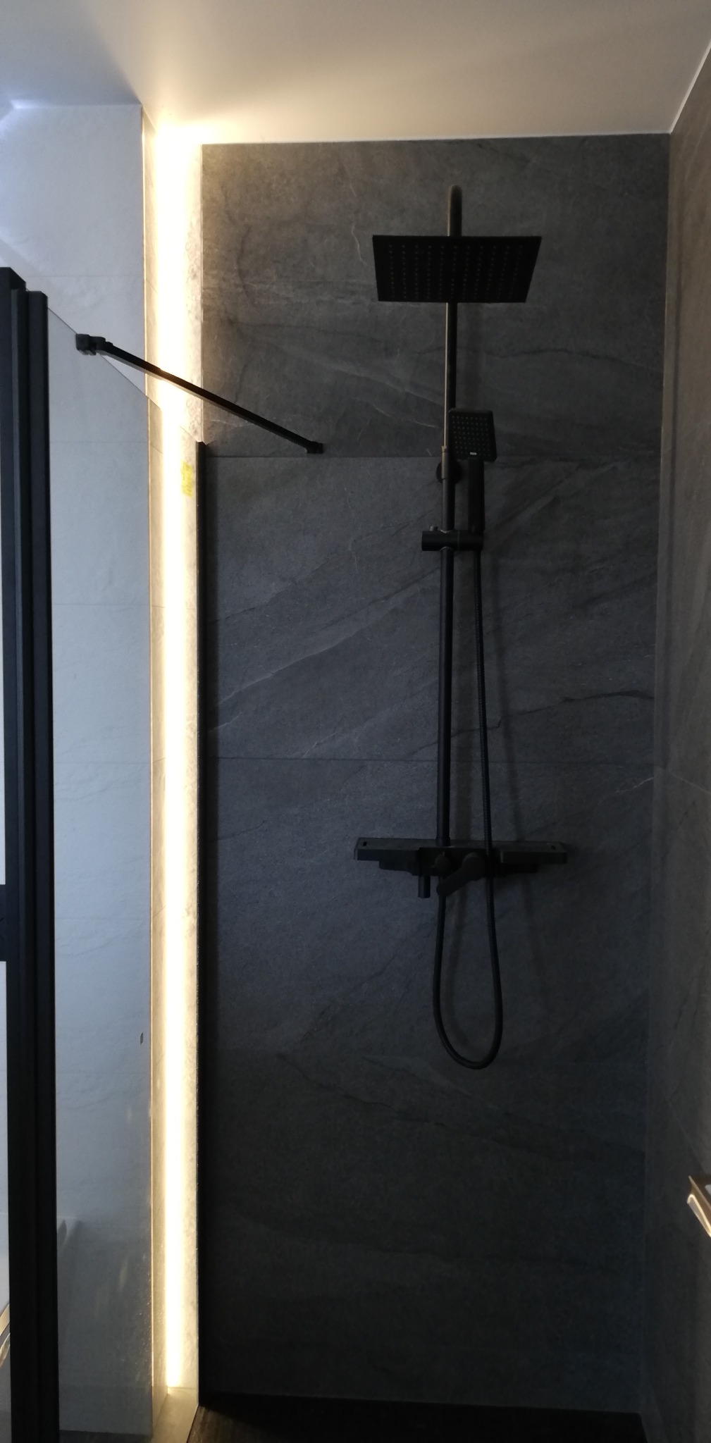 Czarny zestaw prysznicowy z deszczownicą na tle szarych płytek imitujących kamień, oświetlenie LED pionowe przy kabinie prysznicowej.