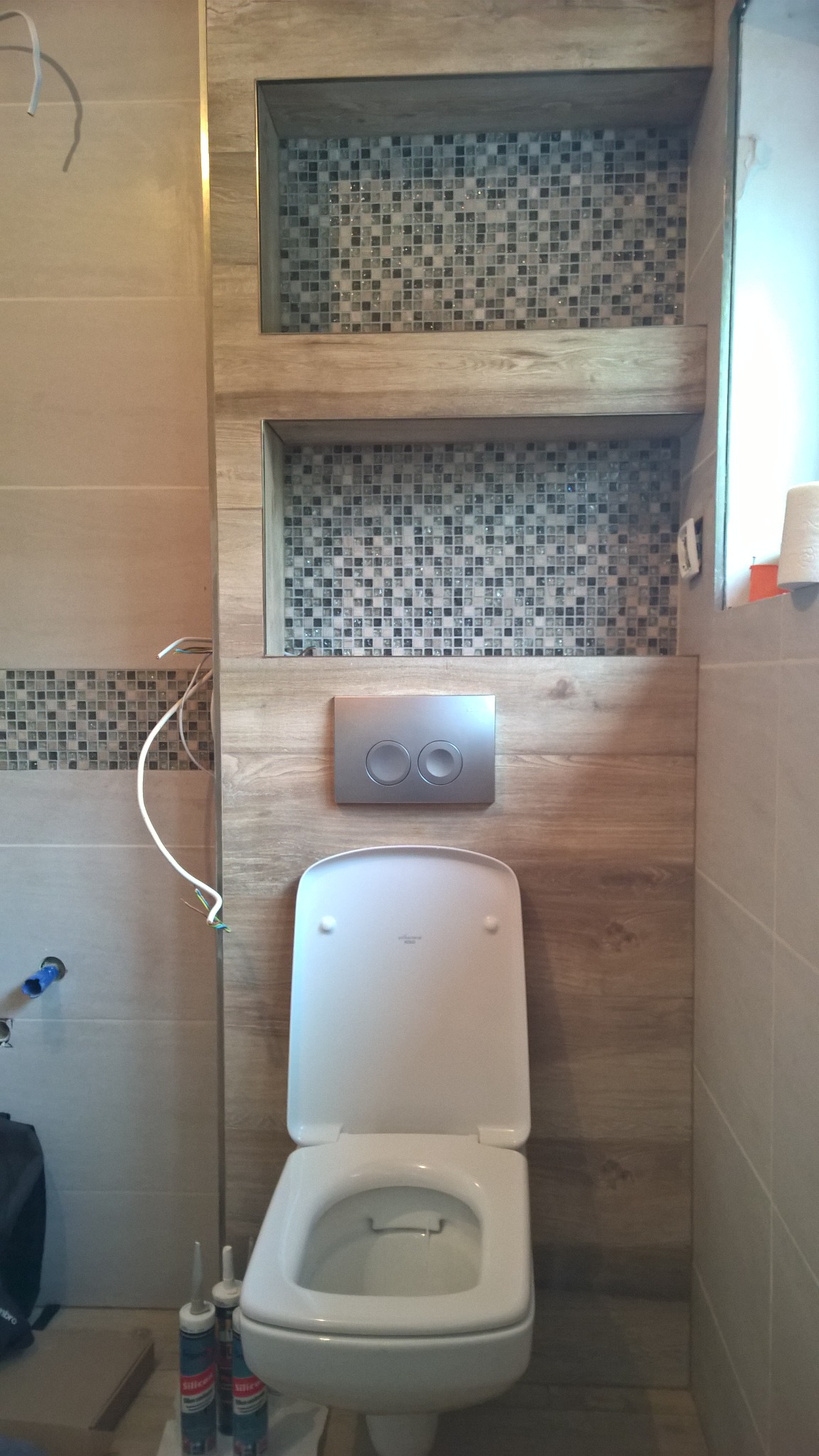 Wykończona łazienka z wiszącą toaletą, drewnopodobnymi panelami ściennymi, mozaiką w półkach i chromowanym przyciskiem spłukiwania, widoczne kable i tuby silikonu.