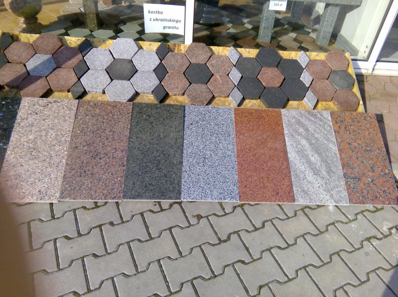 Ekspozycja różnych wzorów i kolorów materiału na blacie: sześciokątne elementy ułożone w kwiatowy wzór oraz prostokątne płyty, prezentowane na zewnątrz na tle chodnika z szarej kostki betonowej.