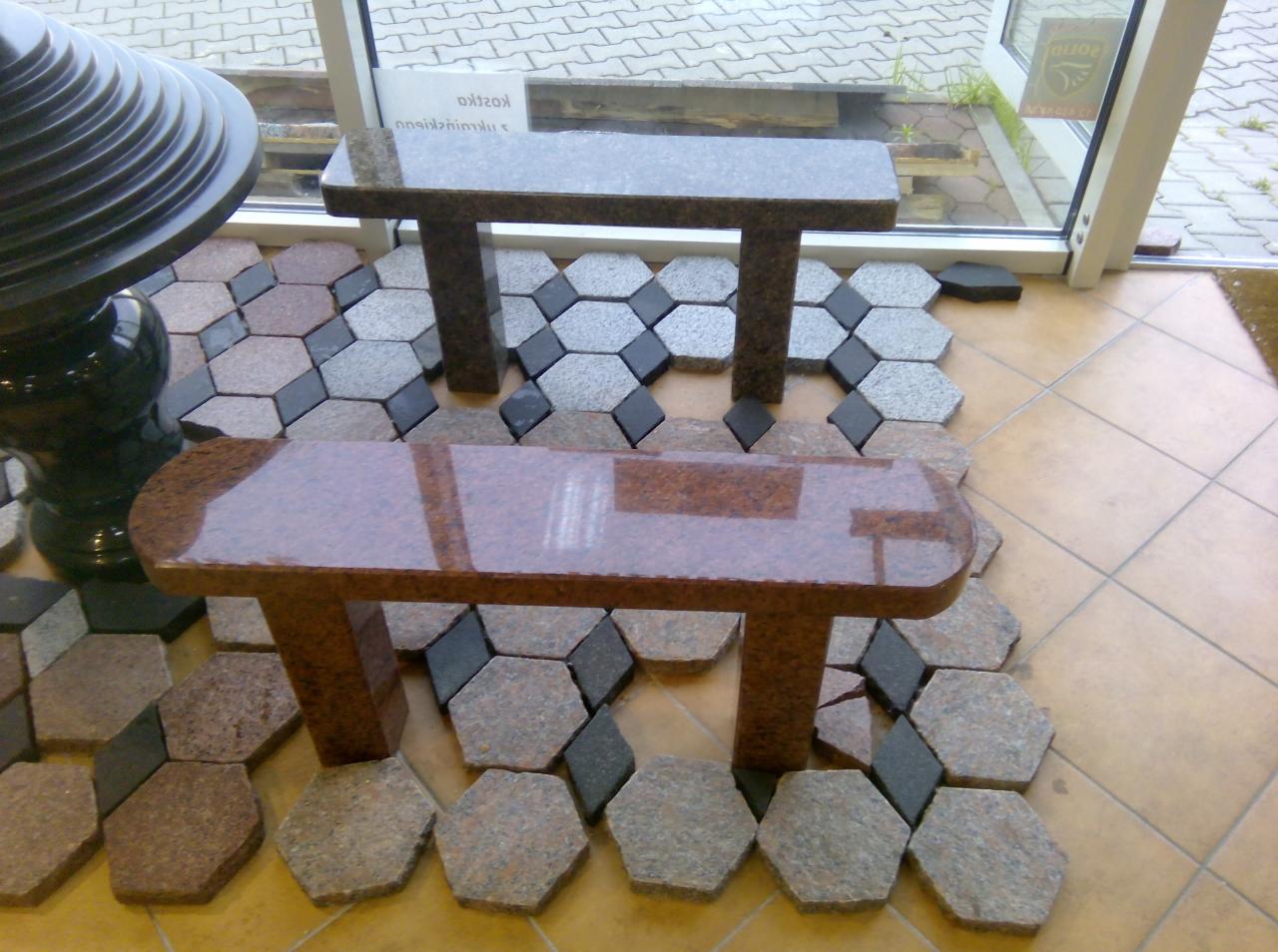 Ekspozycja dwóch granitowych ławek, jedna brązowa, druga szara, ustawionych na podłodze z heksagonalnych elementów w odcieniach szarości, brązu i czerni, obok ciemnozielonej, ozdobnej kolumny.