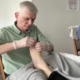 Mężczyzna w zielonej koszuli i rękawiczkach kosmetycznych wykonuje zabieg na stopie klienta leżącego pod szarą pledem, na ścianie wisi schemat refleksologii stóp.