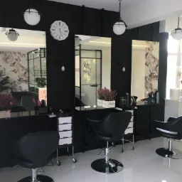 Nowoczesne wnętrze salonu fryzjerskiego z trzema stanowiskami: czarne ściany, duże lustra, designerskie lampy, wygodne fotele fryzjerskie i kwiatowe dekoracje.