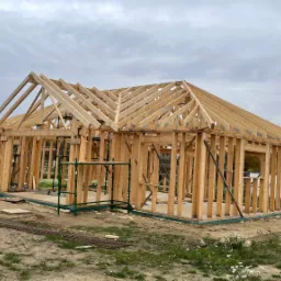 Wznoszenie konstrukcji szkieletowej domu jednorodzinnego, widoczne drewniane elementy ścian i dachu, na zewnątrz rusztowanie i teren budowy w pochmurny dzień.