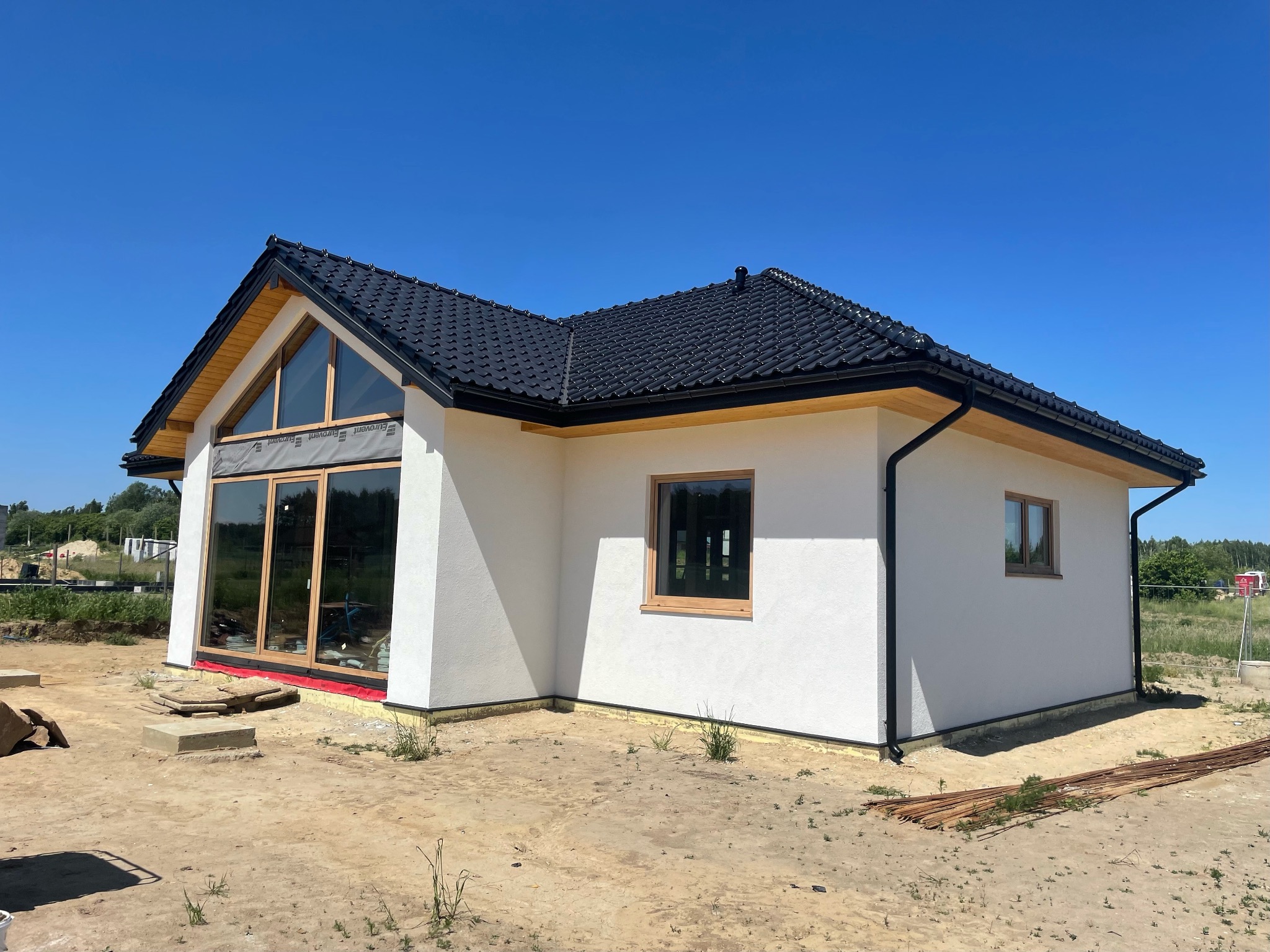 Niedokończony dom szkieletowy z dużymi oknami i czarnym dachem na tle błękitnego nieba, widoczne materiały budowlane wokół.