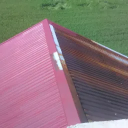Częściowo odnowiony dach z blachy trapezowej: połowa pomalowana na czerwono, druga skorodowana, na tle zielonego pola.