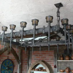 Rustykalny żyrandol z metalu, stylizowany na średniowieczny, z elementami kutymi i imitacją pochodni, zawieszony w pomieszczeniu z ceglaną ścianą i drewnianymi elementami wystroju.