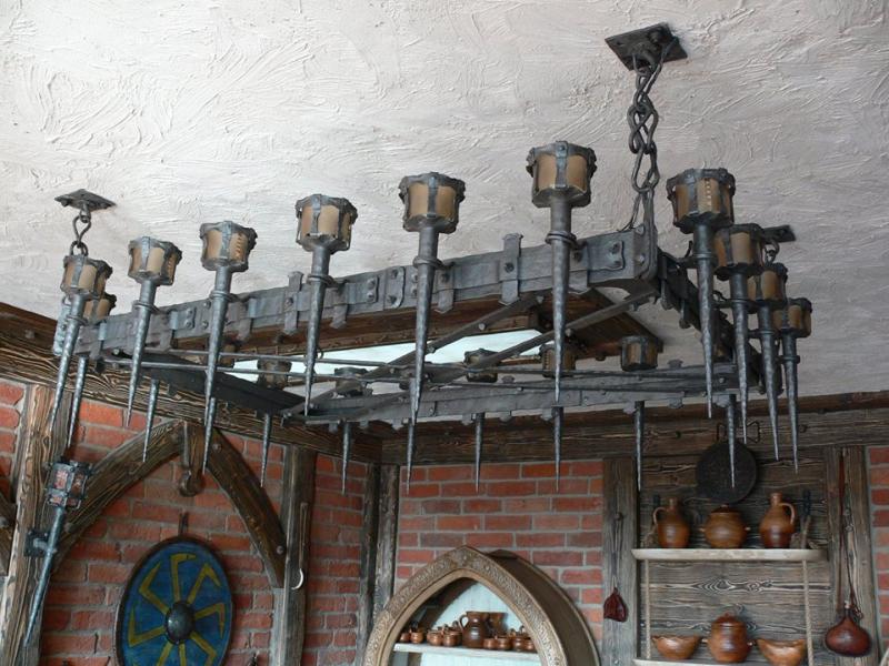 Rustykalny żyrandol z metalu, stylizowany na średniowieczny, z elementami kutymi i imitacją pochodni, zawieszony w pomieszczeniu z ceglaną ścianą i drewnianymi elementami wystroju.
