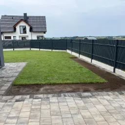Fragment trawnika z nowo ułożonej darni, widoczne łączenia i przygotowane podłoże, obok szara kostka brukowa, w tle nowoczesny dom z ciemną dachówką i ciemnoszarym ogrodzeniem panelowym.