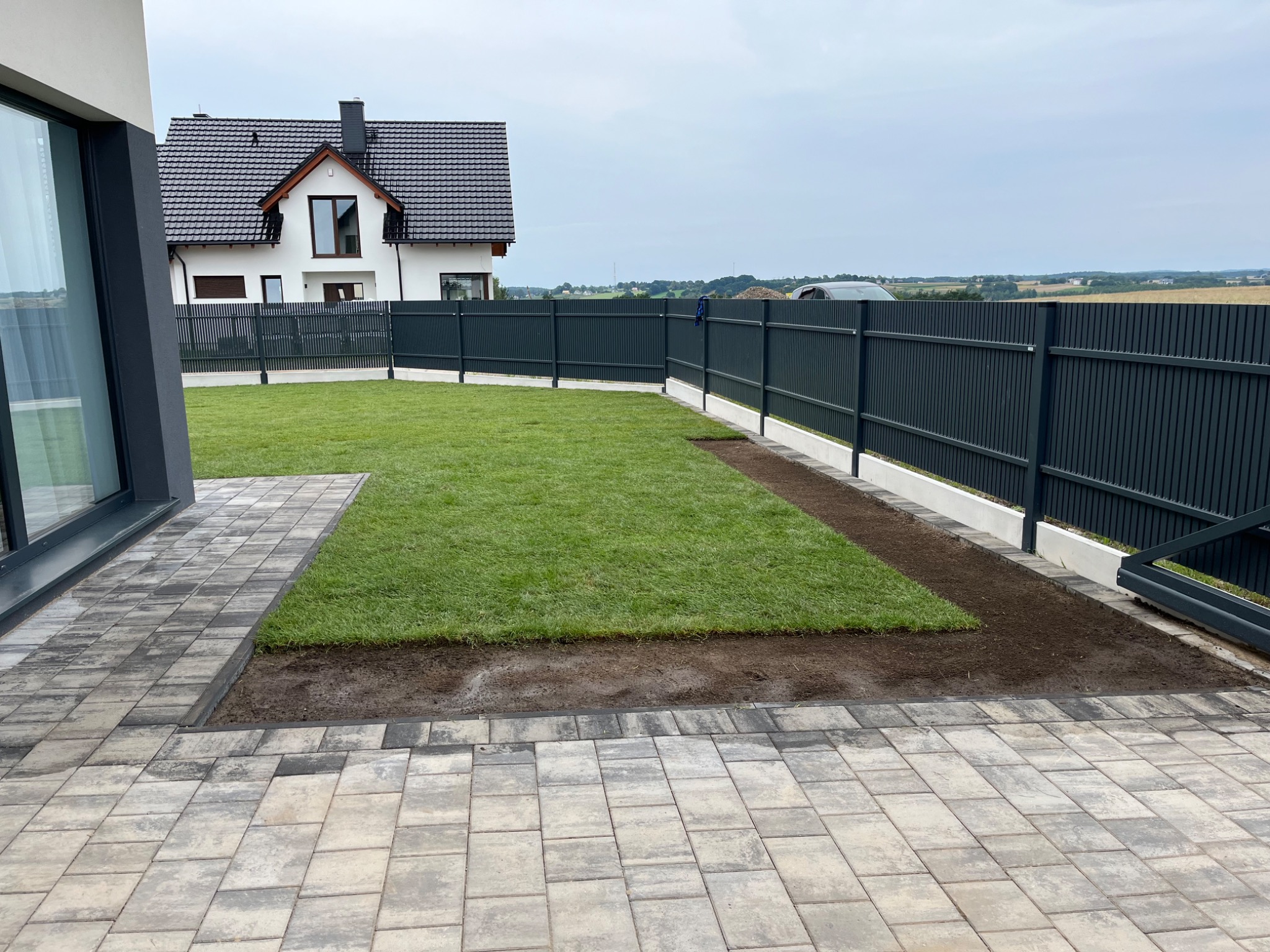 Fragment trawnika z nowo ułożonej darni, widoczne łączenia i przygotowane podłoże, obok szara kostka brukowa, w tle nowoczesny dom z ciemną dachówką i ciemnoszarym ogrodzeniem panelowym.