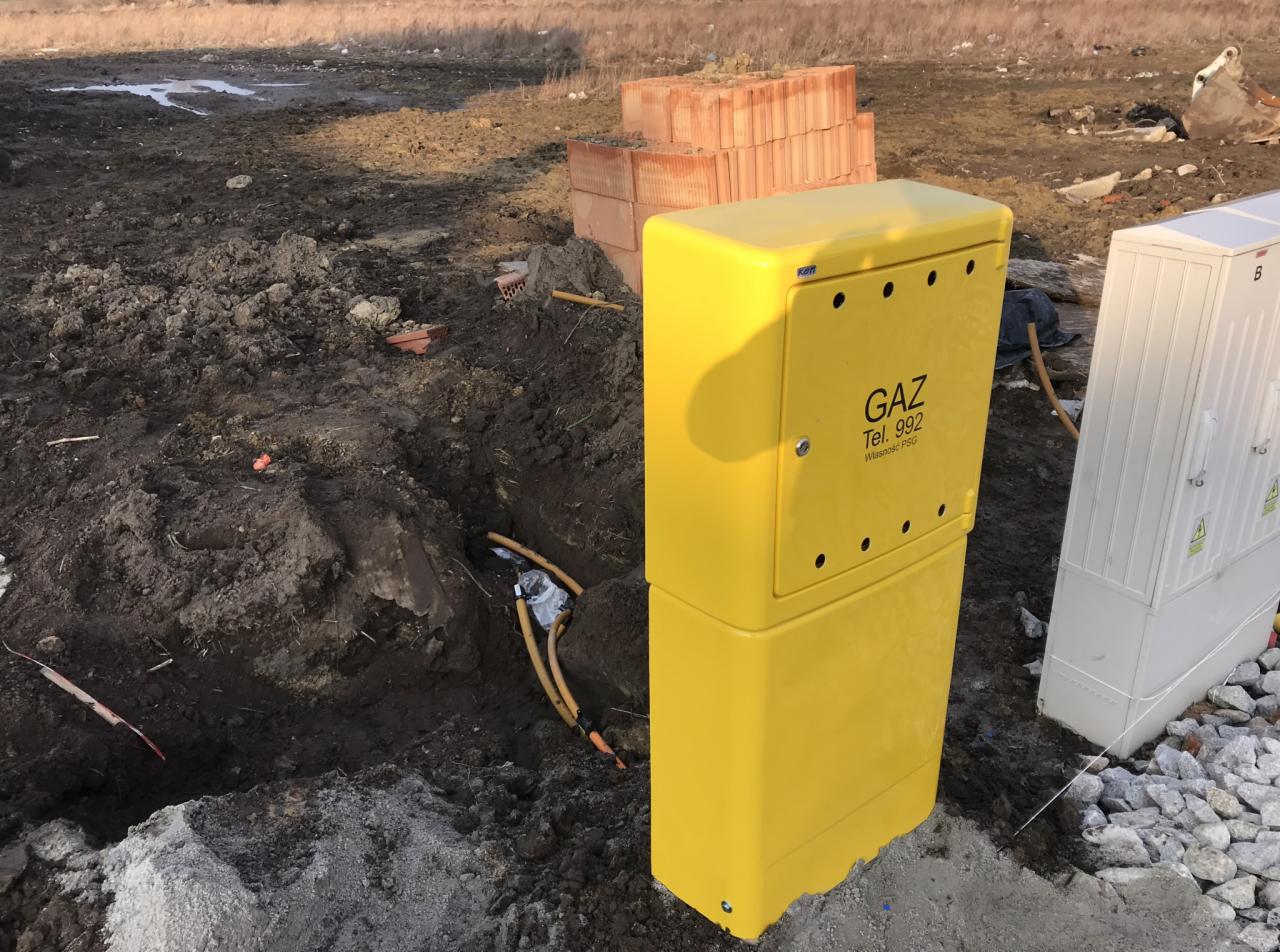 Żółta skrzynka gazowa z napisem 'GAZ', widoczna w wykopie z ułożonymi rurami, obok biała skrzynka i sterta cegieł na placu budowy.