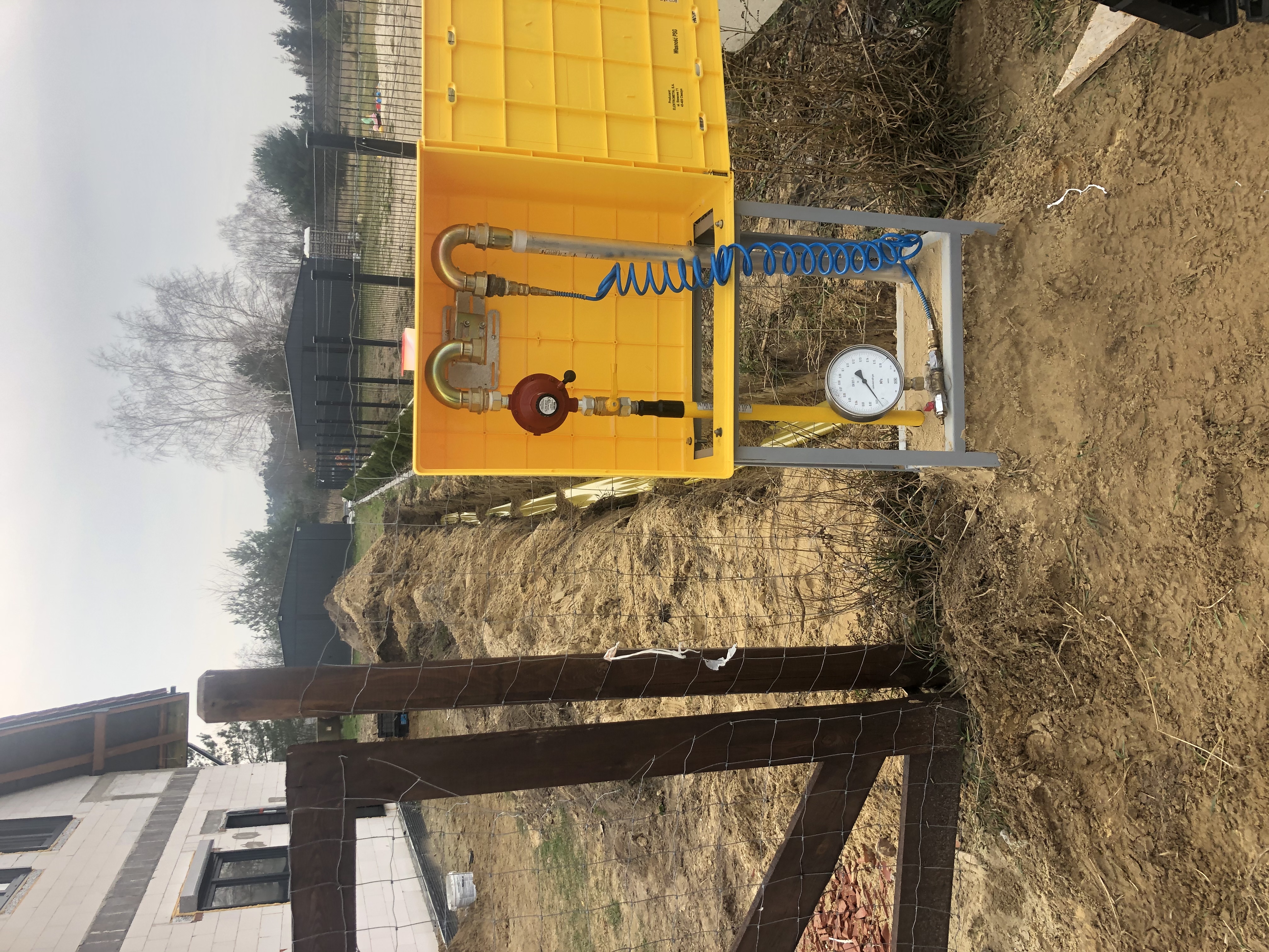 Żółta skrzynka gazowa z reduktorem ciśnienia i manometrem, zamontowana na zewnątrz, widoczna na tle wykopu i ogrodzenia posesji.