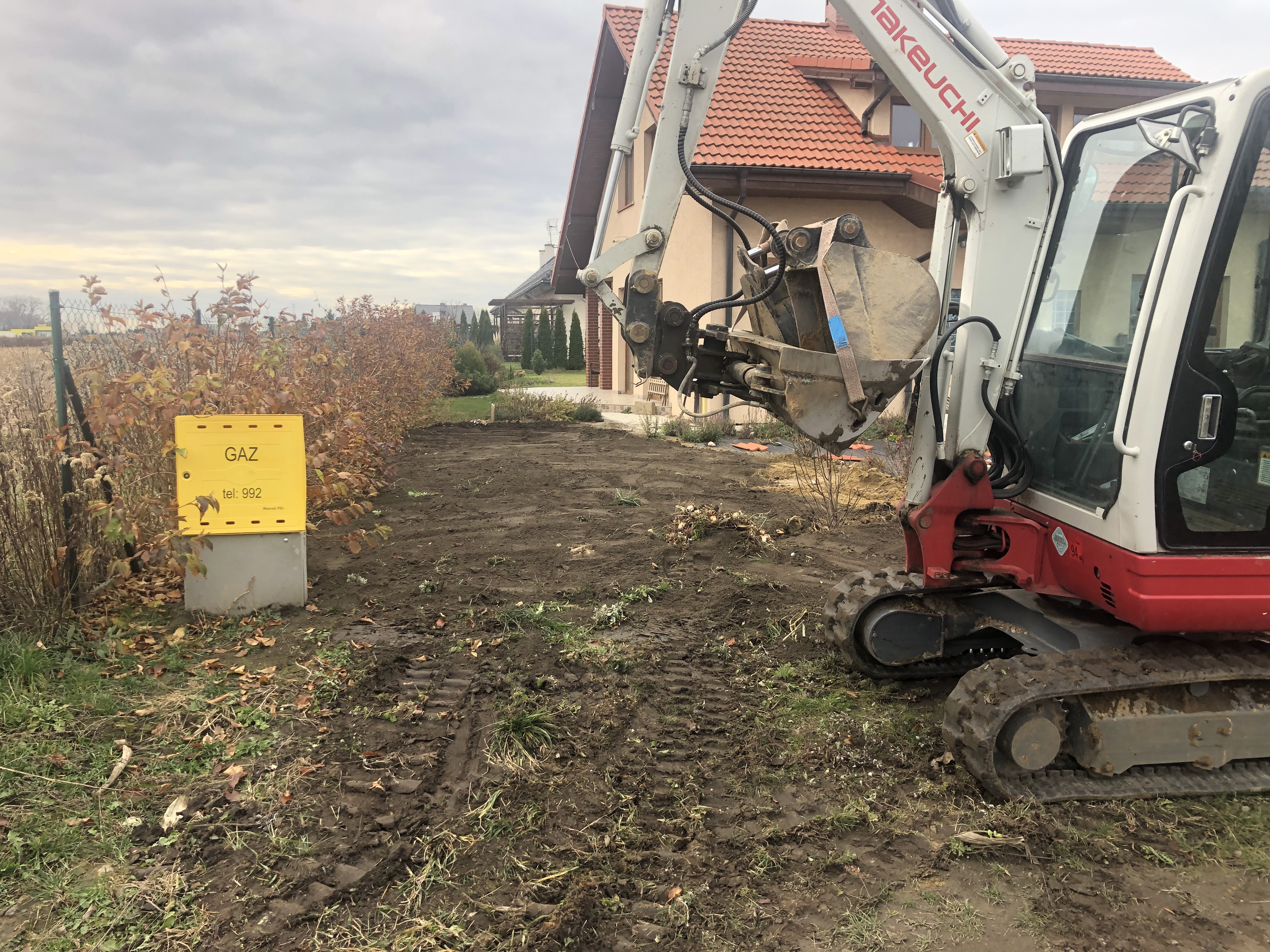 Żółta skrzynka gazowa z numerem telefonu na tle wykopanej ziemi i koparki Takeuchi, przygotowanie terenu pod przyłącze gazowe.