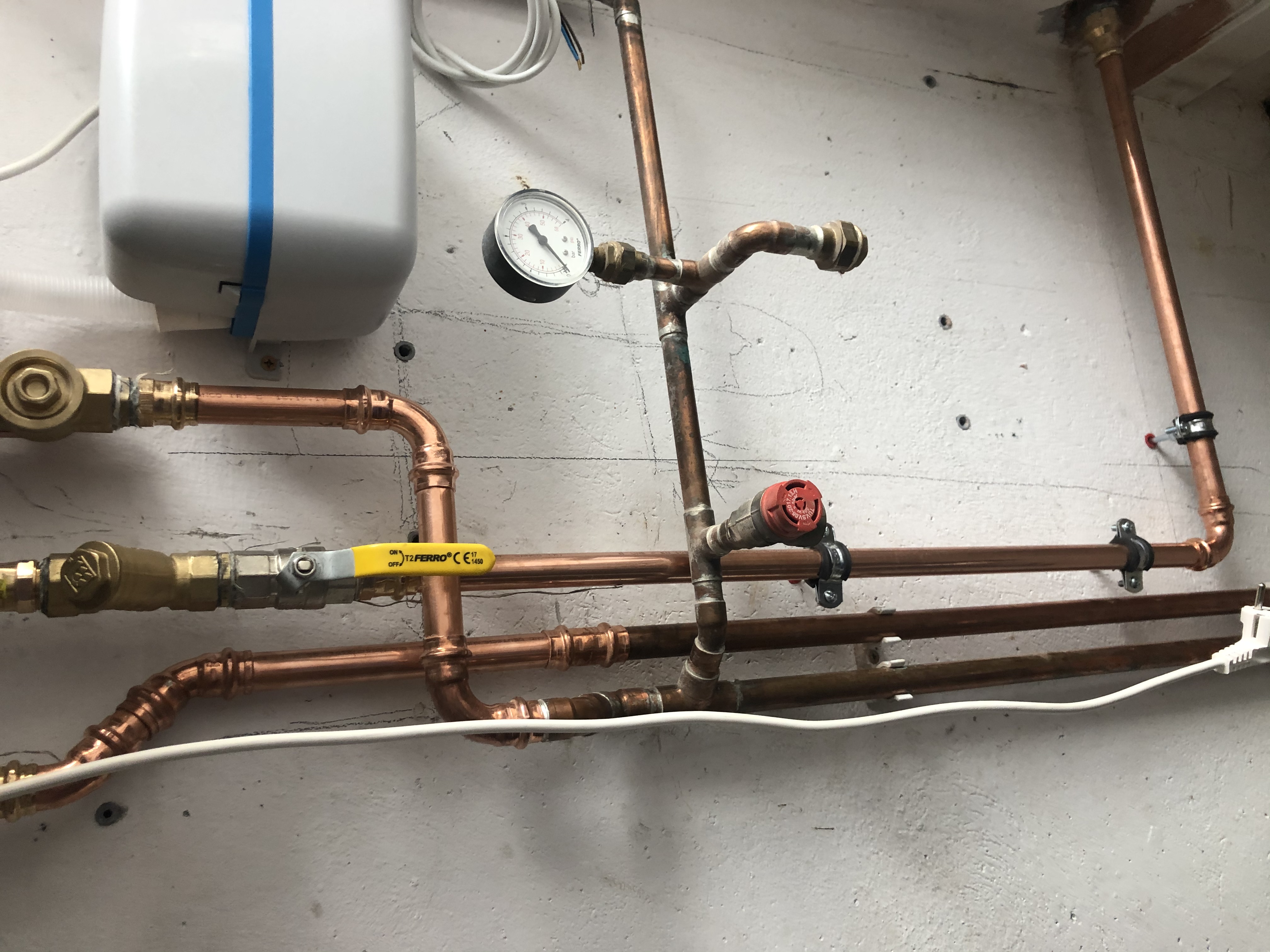Miedziane rury instalacji grzewczej z zaworami, manometrem i białym zasobnikiem na tle szarej, chropowatej ściany.