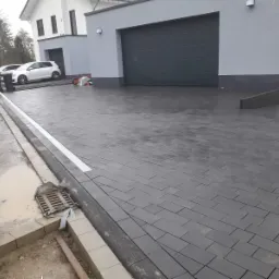 Ułożona, szara kostka brukowa na podjeździe do nowoczesnego domu z garażem, widoczne ślady po budowie i kratka ściekowa przy krawężniku.