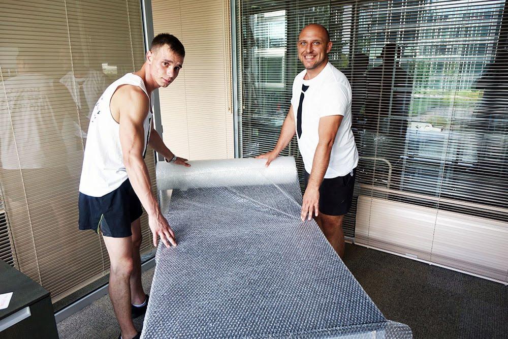 Dwóch mężczyzn owija mebel folią bąbelkową w jasnym pomieszczeniu z żaluzjami.