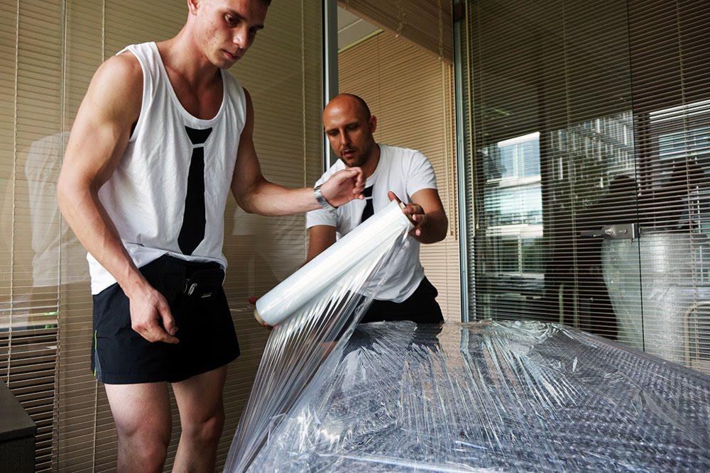 Dwóch pracowników w Warszawie owija folią stretch mebel przed transportem, jeden z nich ma na sobie podkoszulek z nadrukowanym krawatem.
