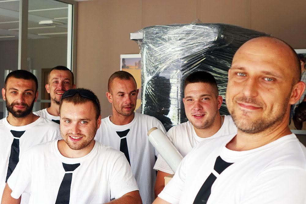 Zespół sześciu mężczyzn w białych koszulkach z czarnymi krawatami pozuje na tle mebla owiniętego folią stretch, sugerując przygotowanie do przeprowadzki.
