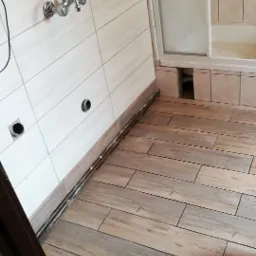 Ukończona podłoga z płytek imitujących drewno w łazience, widoczne przygotowane otwory na rury i wystające rury, ściany wyłożone jasnymi, poziomymi płytkami, fragment kabiny prysznicowej w tle.