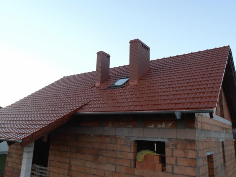 Ceglastoczerwony dach z dwoma kominami i oknem dachowym na niedokończonym budynku z cegły, z zamontowanymi rynnami.