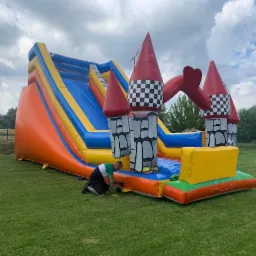 Duży, kolorowy dmuchany zamek ze zjeżdżalnią na trawie, mężczyzna w krótkich spodenkach i koszulce z krótkim rękawem naprawia podstawę zamku.