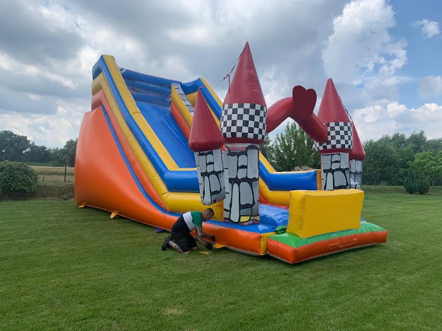 Duży, kolorowy dmuchany zamek ze zjeżdżalnią na trawie, mężczyzna w krótkich spodenkach i koszulce z krótkim rękawem naprawia podstawę zamku.