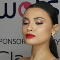 Portret kobiety z zamkniętymi oczami, prezentującej intensywny makijaż oczu w odcieniach brązu i czerwone usta, z widocznymi kolczykami na tle ściany z napisem sponsora.