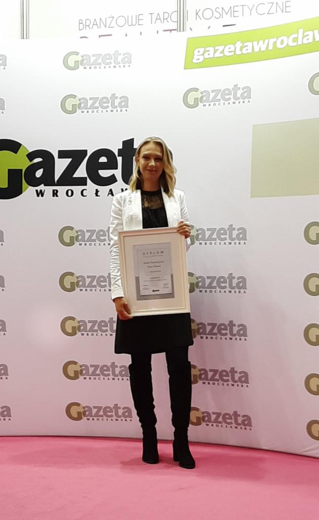 Kobieta w białej marynarce prezentuje dyplom na tle ściany z logotypami Gazety Wrocławskiej podczas branżowych targów kosmetycznych.