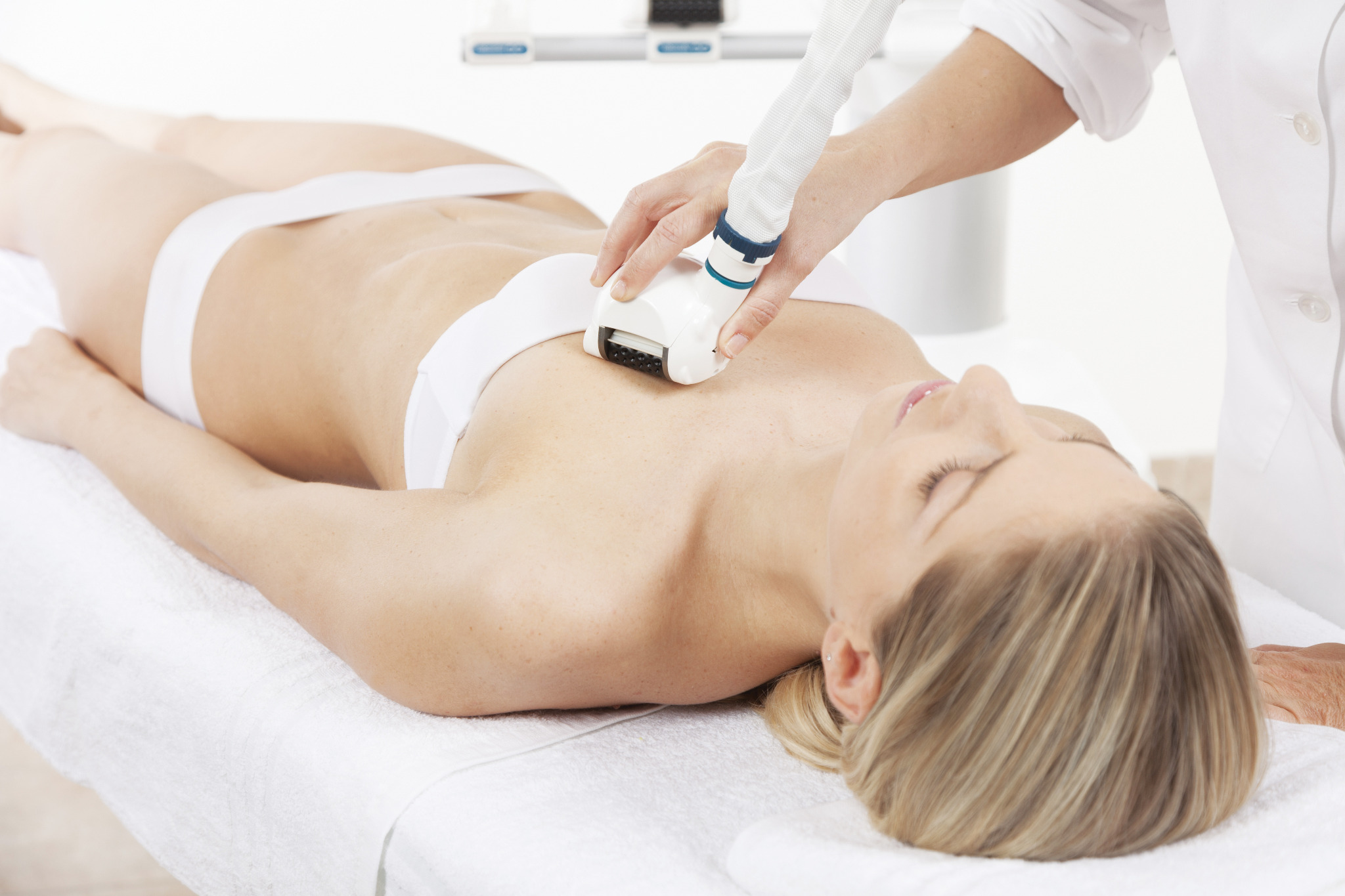 Kobieta leżąca na leżance podczas zabiegu depilacji laserowej klatki piersiowej, widoczny aplikator urządzenia w dłoni pracownika w białym uniformie.