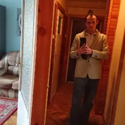 Mężczyzna w beżowej marynarce robi selfie w lustrze w korytarzu z boazerią i drewnianą podłogą w jodełkę.