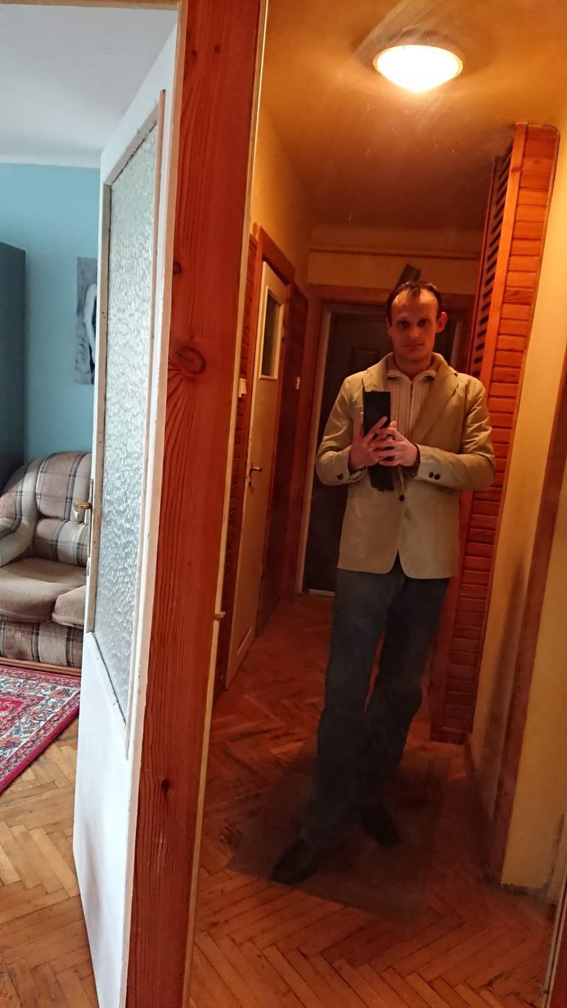 Mężczyzna w beżowej marynarce robi selfie w lustrze w korytarzu z boazerią i drewnianą podłogą w jodełkę.