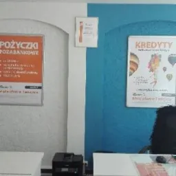 Wnętrze biura z plakatami reklamującymi pożyczki i kredyty na ścianach, biurka i drukarka w tle.