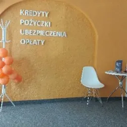 Wnętrze biura z napisem na ścianie 'Kredyty Pożyczki Ubezpieczenia Opłaty', obok wieszak z pomarańczowymi balonami, białe krzesło i mały stolik z ulotkami.