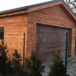 Drewniany garaż z bramą segmentową i małym oknem, otoczony tujami i ogrodzeniem z siatki.