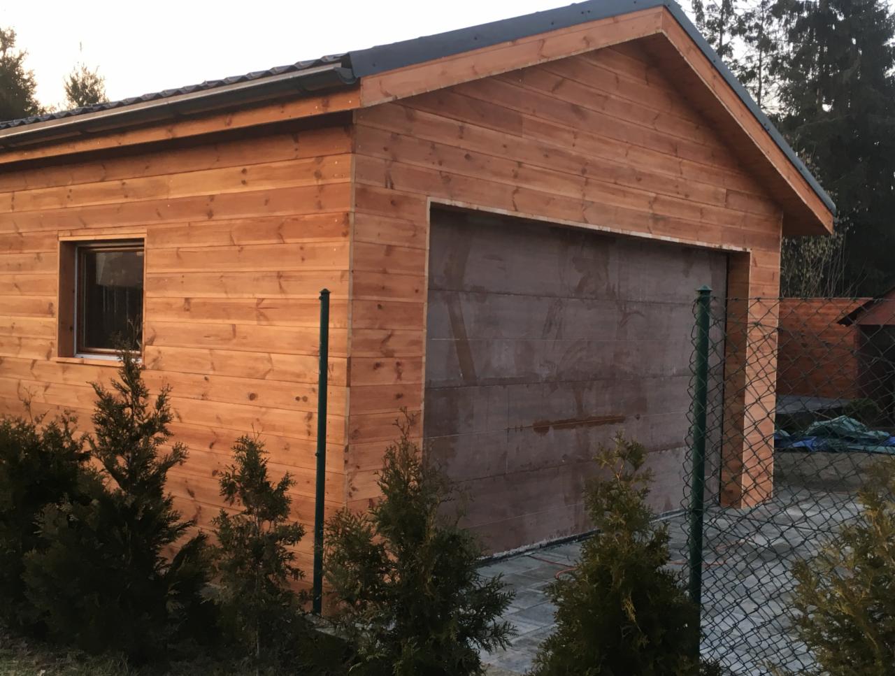 Drewniany garaż z bramą segmentową i małym oknem, otoczony tujami i ogrodzeniem z siatki.