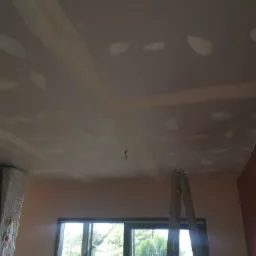 Sufit z płyt kartonowo-gipsowych w trakcie szpachlowania, widoczne łączenia i miejsca po wkrętach pokryte gładzią, drabina oparta o ścianę, okno z widokiem na zieleń w tle.