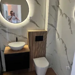 Nowoczesna łazienka z umywalką nablatową na drewnianym blacie, toaletą typu Rimless i okrągłym lustrem z podświetleniem LED, odbijającym fotografa.