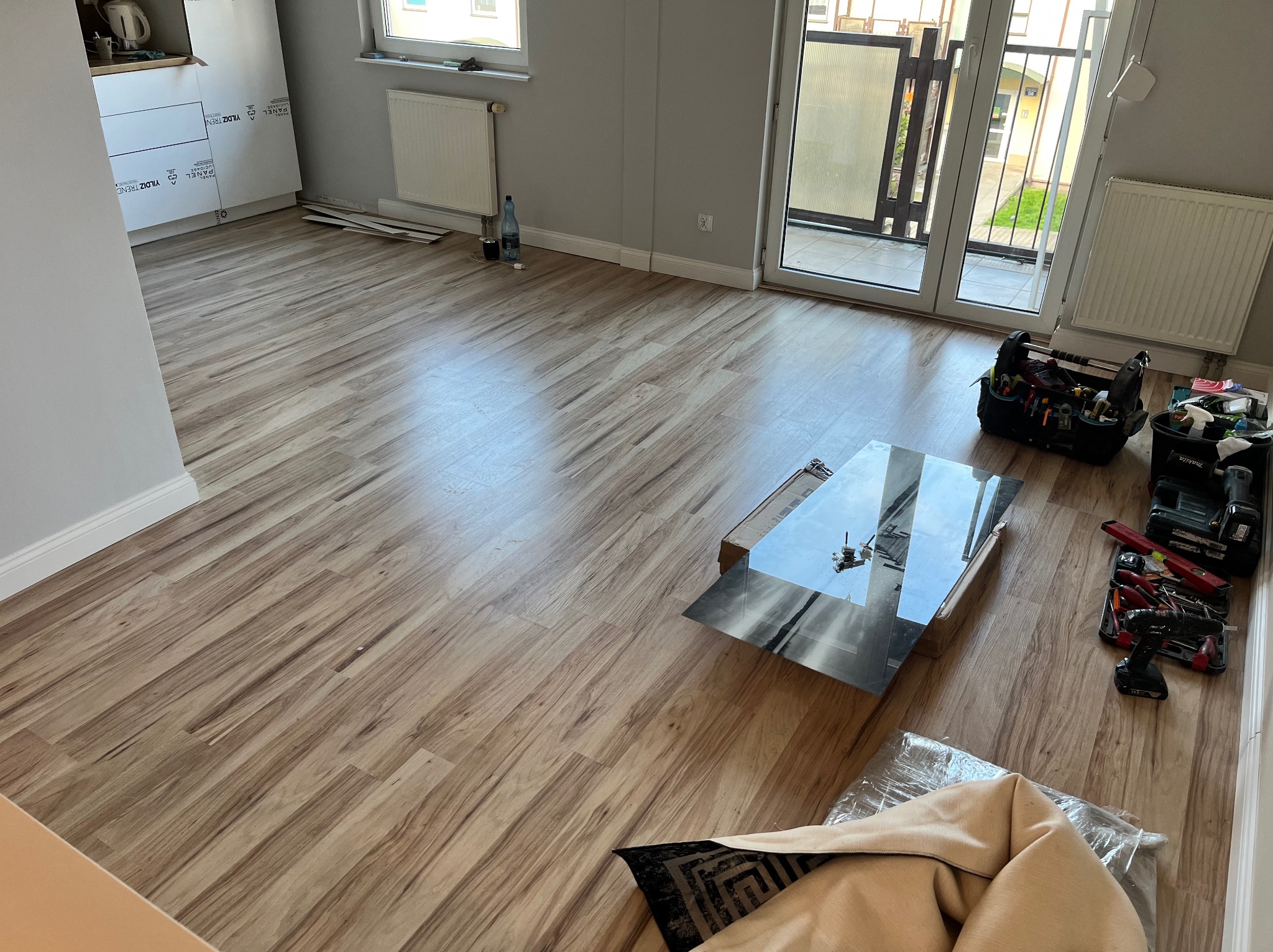 Podłoga wyłożona panelami imitującymi drewno w trakcie montażu, widoczne narzędzia, skrzynka narzędziowa, fragment lustrzanego panelu i materiał zabezpieczający.