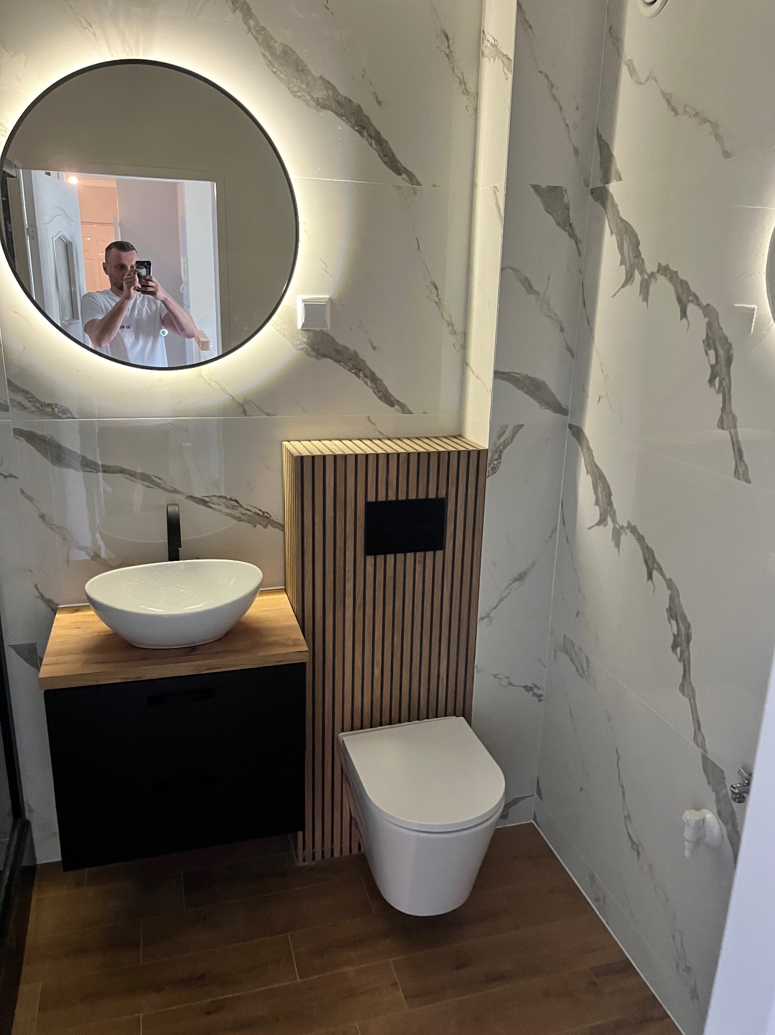 Nowoczesna łazienka z umywalką nablatową na drewnianym blacie, toaletą typu Rimless i okrągłym lustrem z podświetleniem LED, odbijającym fotografa.
