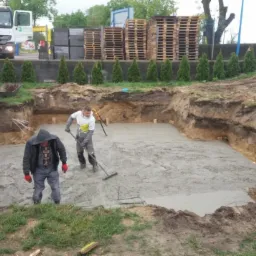 Dwóch pracowników w roboczych ubraniach rozprowadza mokry beton w wykopie pod fundamenty, w tle widoczny samochód ciężarowy i ułożone palety.