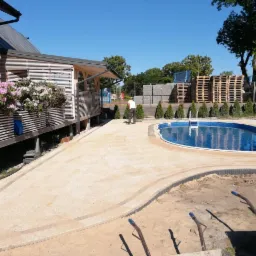 Nowoczesny dom z drewnianym tarasem i kwiatami, obok basen z wykończoną kostką obrzeżową, w tle palety z kostką brukową i pracownik.